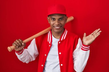 Beyzbol oynayan genç İspanyol adam elinde beyzbol sopasıyla neşeli bir şekilde sunum yapıyor ve avuç içiyle kameraya bakıyor.. 