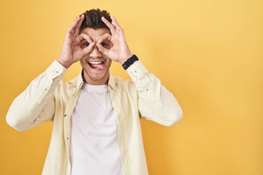 Sarı arka planda dikilen İspanyol genç adam dürbün gibi jest yapıyor. Dürbün dilini çıkarıyor, gözler parmaklarından bakıyor. çılgın ifade. 