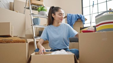 Young beautiful hispanic woman unpacking cardboard box at new home