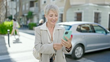 Orta yaşlı sarışın kadın sokakta akıllı telefon kullanarak gülümsüyor.