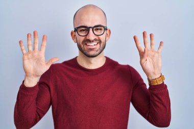 Beyaz arka planda duran sakallı genç ve kel bir adam gözlük takıyor ve dokuz numaralı parmaklarıyla işaret ederken kendinden emin ve mutlu gülümsüyor.. 