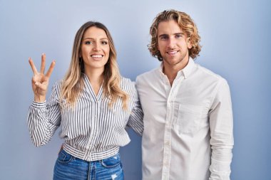 Mavi arka planda duran genç çift üç numaralı parmaklarını gösterip işaret ederken kendinden emin ve mutlu gülümsüyor.. 