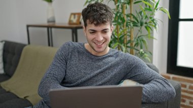 Genç İspanyol adam evdeki koltukta dizüstü bilgisayar kullanıyor.