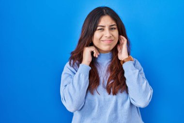 Mavi arka planda dikilen Latin kökenli genç bir kadın yüksek sesli müzik sesi için parmaklarıyla kulaklarını kapatıyor. sağır kavramı. 