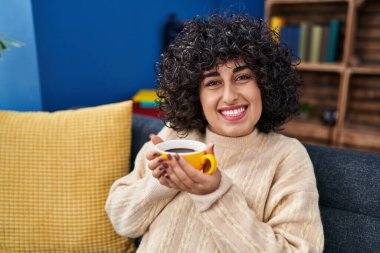 Orta Doğu 'lu genç bir kadın evdeki kanepede kahve içiyor.