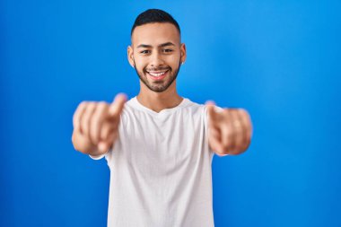 Young hispanic man standing over blue background pointing to you and the camera with fingers, smiling positive and cheerful 