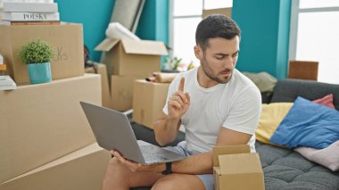 Young hispanic man using laptop sitting on sofa at new home