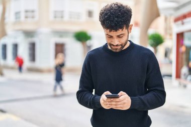 Genç Arap adam sokakta akıllı telefon kullanarak kendinden emin gülümsüyor.