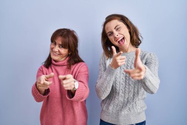 Anne ve kızı mavi arka planda durmuş mutlu ve komik yüzlü parmaklarını kameraya doğrultuyorlar. iyi enerji ve titreşimler. 