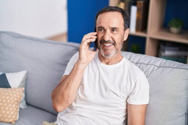 Orta yaşlı bir adam akıllı telefonda konuşuyor. Evdeki kanepede oturuyor.