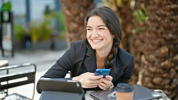 コーヒーショップのテラスで笑顔のスマートフォンを使用して若い美しいヒスパニック系女性のビジネスワーカー — ストック写真