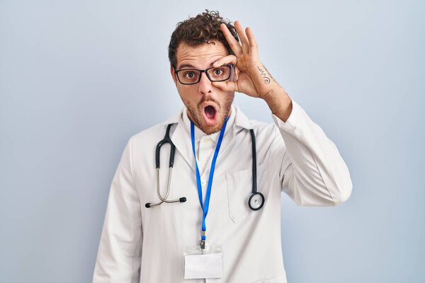 Young hispanic man wearing doctor uniform and stethoscope doing ok gesture shocked with surprised face, eye looking through fingers. unbelieving expression. 