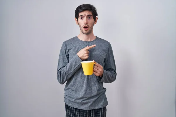 Ung Latinamerikan Man Bär Pyjamas Dricka Kopp Kaffe Förvånad Pekar — Stockfoto