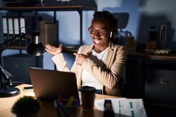 Beautiful black woman working at the office at night amazed and smiling to the camera while presenting with hand and pointing with finger.
