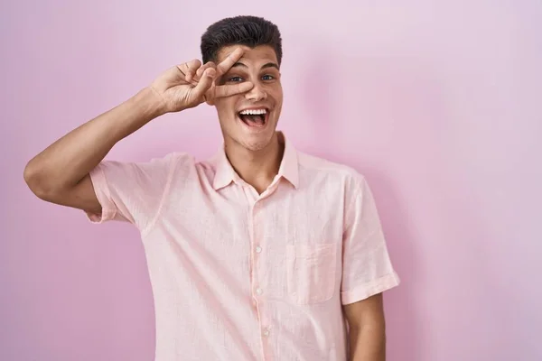 ピンクの背景の上に立つ若いヒスパニック系の男は 顔の上に指で平和のシンボルをやって 勝利を示す明るい笑顔 — ストック写真