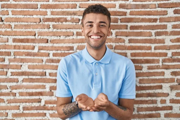 手の手のひらで一緒に受信またはジェスチャーを与える笑みを浮かべてレンガの壁の上に立ってブラジルの若い男 保持し保護する — ストック写真