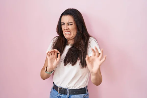 Young brunette woman standing over pink background disgusted expression, displeased and fearful doing disgust face because aversion reaction.