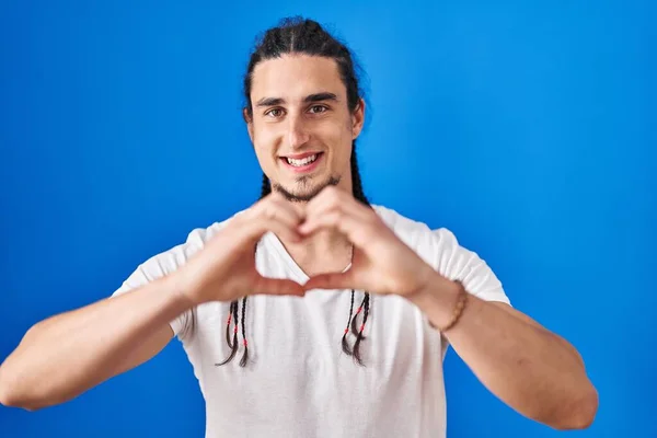 一个长头发的西班牙男人站在蓝色的背景上 微笑着 用双手做着心形符号 浪漫的概念 — 图库照片