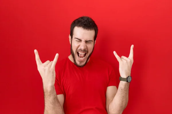 手を上げて岩のシンボルをするクレイジーな表情で叫んでカジュアルな赤いTシャツを着た若いヒスパニック系の男 音楽スター 重い音楽の概念は — ストック写真