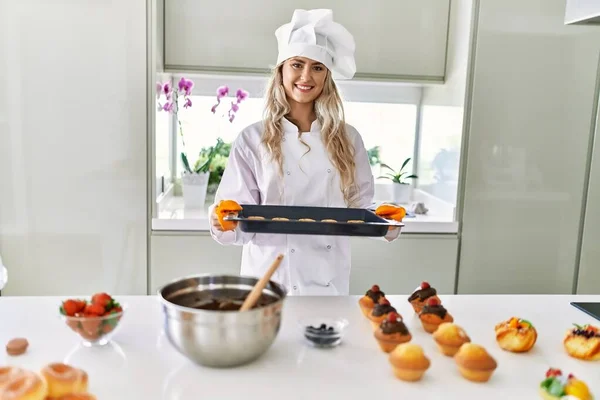 stock image Young woman wearing cook uniform holding oven tray with cookies at kitchen