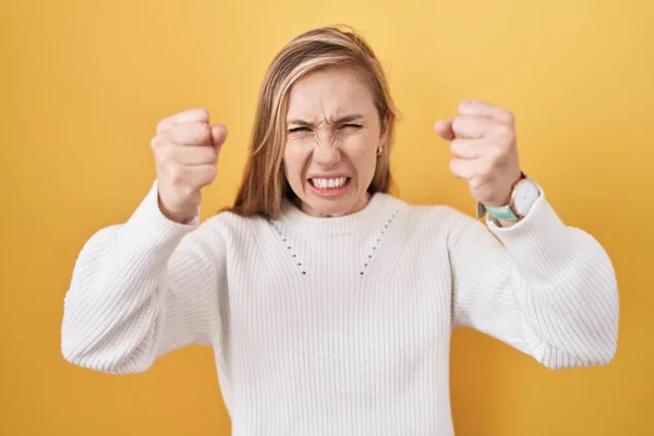 怒って怒った黄色の背景に白いセーターを着た若い白人女性が怒りながら叫びながら拳をフラストレーションと激怒させます 怒りと攻撃的な考え方 — ストック写真