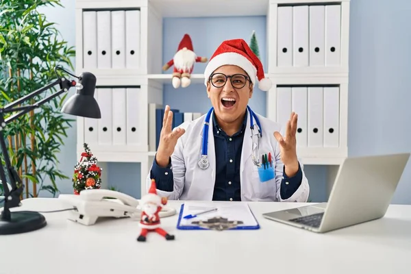 Hispanic young doctor man at the clinic on christmas crazy and mad shouting and yelling with aggressive expression and arms raised. frustration concept.