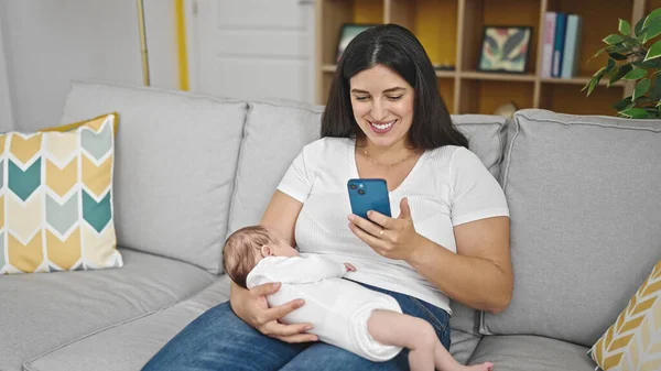 自宅でスマートフォンを使用して赤ちゃんを抱えているソファーに座っている母と赤ちゃんの娘 — ストック写真
