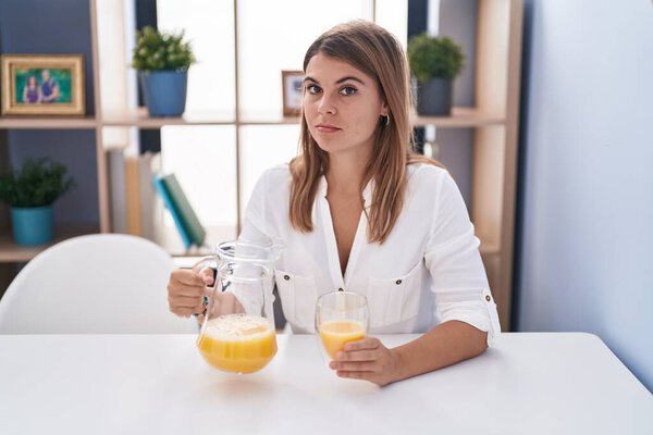 Young hispanic woman drinking glass of orange juice relaxed with serious expression on face. simple and natural looking at the camera. 