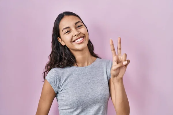 ピンクの背景にカジュアルなTシャツを着た若いブラジル人女性が 勝利サインをしている指を示すカメラを見て笑っています — ストック写真