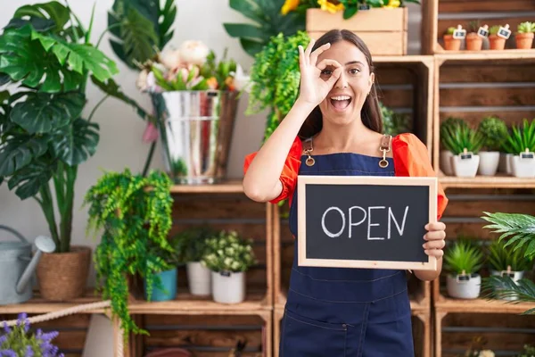 stock image Young hispanic woman working at florist holding open sign smiling happy doing ok sign with hand on eye looking through fingers 