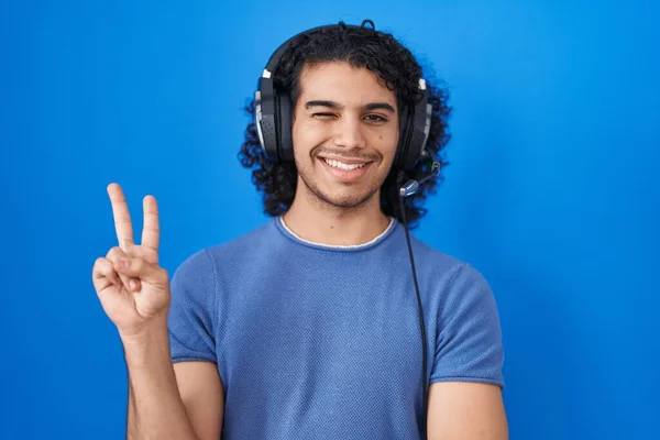 カメラでの勝利サインを行う幸せな顔のウィンクで笑顔ヘッドフォンを使用して音楽を聞いて巻き毛を持つヒスパニック系の男 — ストック写真