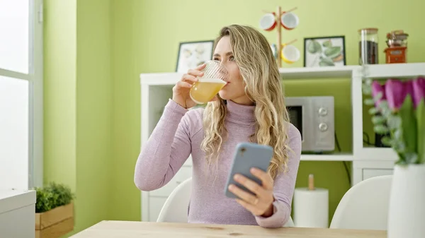若いですブロンド女性飲むガラスのオレンジジュース使用スマートフォンでダイニングルーム — ストック写真
