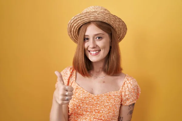 年轻的红头发女人站在黄色的背景上 戴着夏帽 手拿着快乐的大拇指做手势 赞成的表情看着相机显示的成功 — 图库照片