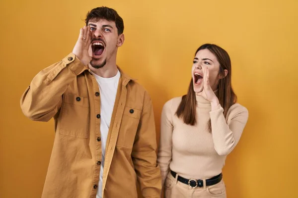 若いヒスパニック系のカップルは黄色の背景に叫んで立っていると大声で口の上に手で横に叫んでいる コミュニケーションの概念 — ストック写真