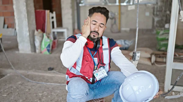 Young latin man builder tired sitting on floor at construction site
