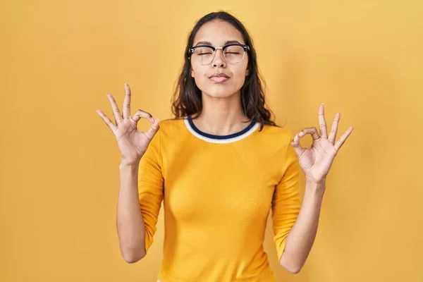 黄色の背景に眼鏡をかけた若いブラジル人女性がリラックスし 目を閉じて指で瞑想ジェスチャーを行う笑顔 ヨガのコンセプト — ストック写真