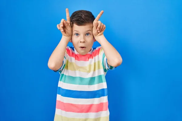 年轻的高加索小孩站在蓝色的背景上做着滑稽的动作 手指头对头像公牛的角 — 图库照片