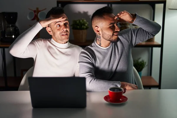 Homosexual couple using computer laptop very happy and smiling looking far away with hand over head. searching concept.