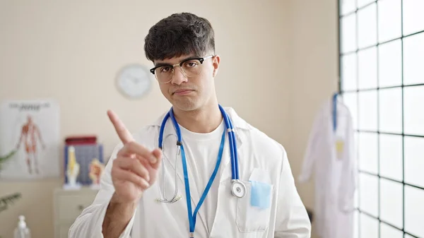 Young hispanic man doctor standing with serious expression saying no with finger at clinic