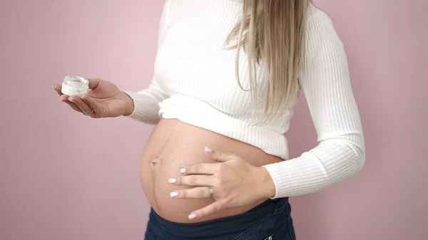 年轻孕妇在腹部涂护肤霜 并涂在孤立的粉色背景上 — 图库照片