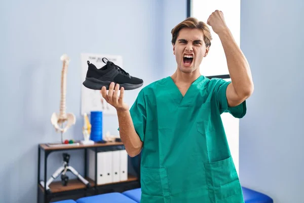 Młody Człowiek Pracujący Klinice Fizjoterapii Trzymający Buty Zirytowany Sfrustrowany Krzykiem — Zdjęcie stockowe