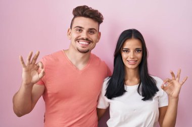 Pembe arka planda durup gülümseyen genç İspanyol çift el ve parmaklarıyla 
