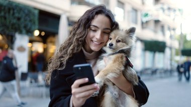 Köpeğiyle gülümseyen genç İspanyol kadın. Sokakta akıllı telefon kullanıyor.
