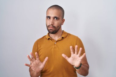 Hispanic man with beard standing over white background afraid and terrified with fear expression stop gesture with hands, shouting in shock. panic concept. 