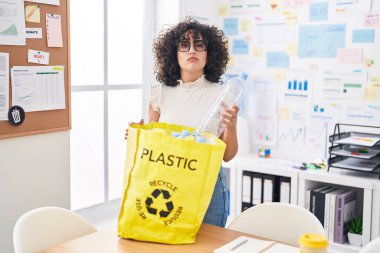 Orta Doğulu genç bir kadın elinde plastik şişelerle geri dönüşüm torbası tutuyor şüpheci ve gergin, sorun yüzünden suratını asıyor. negatif kişi. 