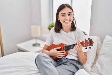 Young hispanic woman playing ukelele sitting on bed at bedroom
