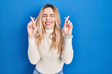 Young caucasian woman standing over blue background gesturing finger crossed smiling with hope and eyes closed. luck and superstitious concept. 