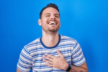 Mavi arka planda dikilen Latin asıllı genç adam gülümseyerek ve kahkahalarla gülüyor çünkü elleri vücutta olan komik çılgın bir şaka.. 