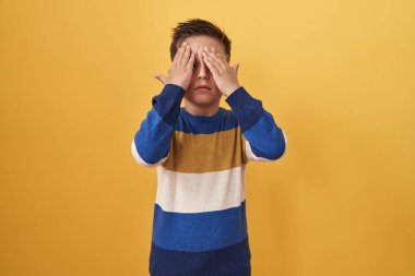 Sarı arka planda duran küçük İspanyol çocuk, elleri başının üstünde, stresten baş ağrısı çekiyor. migren ağrısı. 