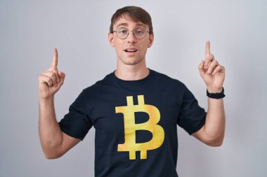 Beyaz, sarışın, bitcoin tişört giyen, şaşkın, şaşkın, parmaklarıyla işaret eden ve kollarını kaldıran bir adam.. 